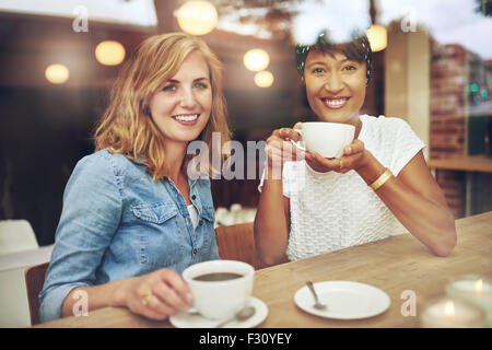 Attraktive junge Multi ethnischen Freundinnen sitzen zusammen an einem Tisch in einem Café genießen Kaffee Lächeln glücklich bei der Zertifizierungsstelle Stockfoto