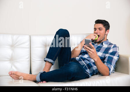 Porträt von ein gut aussehender Mann mit Smartphone und Essen Apfel auf dem sofa Stockfoto