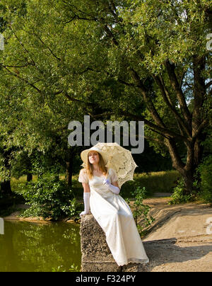 Junge Dame in weiß historische Kleid, Hut und Sonnenschirm sitzen in der Nähe von See im Park. Stockfoto