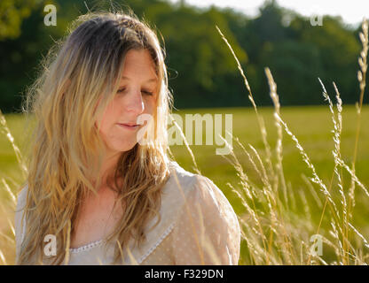 Eine Hintergrundbeleuchtung Porträt einer Frau (im Alter von 25-30) trägt ein weißes Top, saß in einem Feld lange Gras an einem Sommerabend Stockfoto