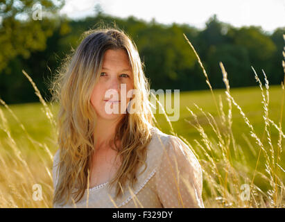 Eine Hintergrundbeleuchtung Porträt einer Frau (im Alter von 25-30) trägt ein weißes Top, saß in einem Feld lange Gras an einem Sommerabend Stockfoto