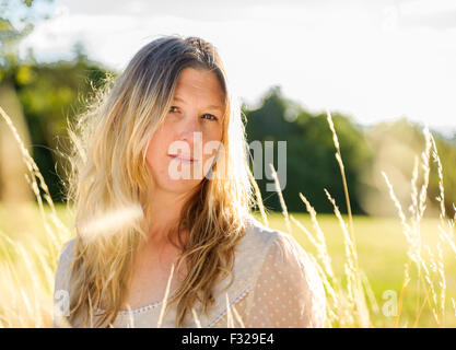 Eine Hintergrundbeleuchtung Porträt einer Frau (im Alter von 25-30) trägt ein weißes Top, saß in einem Feld lange Gras an einem Sommerabend Stockfoto