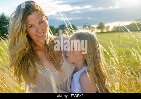 Eine Hintergrundbeleuchtung Porträt einer Frau (Alter 25-30) mit ihrer Tochter (8 Jahre) saß in einem Feld lange Gras auf einen Sommerabend Stockfoto