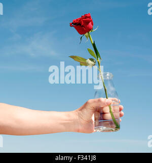 Closeup von einem kaukasischen Jüngling mit einer Glasflasche mit einer roten Rose darin, gegen den blauen Himmel Stockfoto