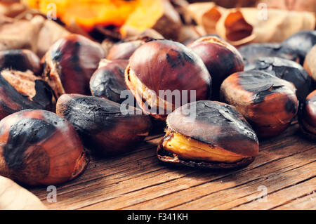 Nahaufnahme von einigen gerösteten Kastanien auf einem rustikalen Holztisch mit einigen gerösteten Süßkartoffeln und Herbstlaub im Hintergrund Stockfoto