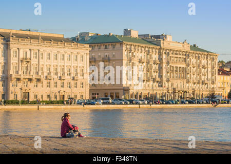 Hafen von Triest Italien, Blick bei Sonnenuntergang auf eine junge Frau, die allein auf dem riesigen Steinsteg (Molo Audace) im Hafen von Triest sitzt Stockfoto