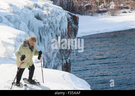 Ältere Frau mit Schneeschuhen bewegt sich über eine verschneite Bank hoch über Lake Superior im Winter, Tettegoche, MN, USA Stockfoto