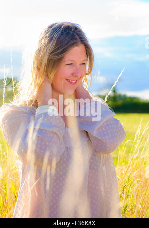 Eine Hintergrundbeleuchtung Porträt einer Frau (im Alter von 25-30) trägt ein weißes Top, Stand in einem Feld lange Gras an einem Sommerabend Stockfoto