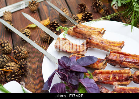 Drei Spieße mit gegrilltem Rippen liegen auf einem weißen Teller mit Cherry-Tomaten, Zwiebeln und Kräutern Stockfoto