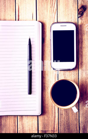 Weiße, Handy, Kugelschreiber, Tasse Kaffee und Notebook auf Holztisch. Arbeitsbereich. Instagram Vintage Bild. Stockfoto
