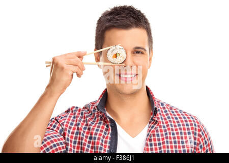 Young guy ein Stück Sushi mit chinesischen Stäbchen vor seinem rechten Auge und schaut in die Kamera Stockfoto