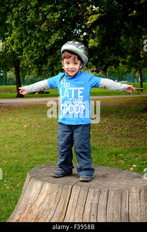 Ein kleiner Junge, das Tragen eines Helms beim Radfahren steht auf einem Baumstumpf im Park. Stockfoto