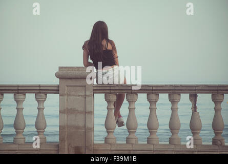 Rückansicht der ein junges Mädchen sitzt auf einer Mauer mit Blick aufs Meer Stockfoto