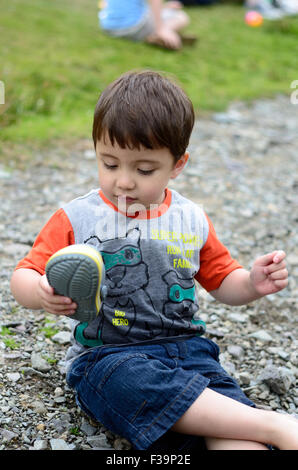 Ein kleiner Junge sitzt auf dem Boden auf einem Stein aus seinem Schuh zu entfernen. Stockfoto