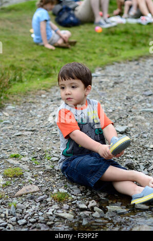 Ein kleiner Junge sitzt im Boden am Rande eines Baches, ein Stein aus seinem Schuh zu bekommen. Stockfoto