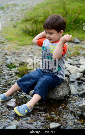 Ein kleiner Junge sitzt auf einem Stein in der Mitte einen stream Stockfoto