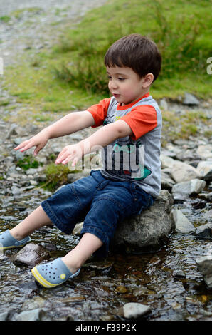 Ein kleiner Junge sitzt auf einem Stein in der Mitte einen stream Stockfoto