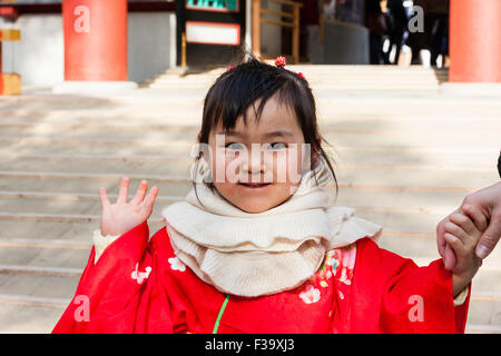 Japanisches Kind, Mädchen, 3-4 Jahre alt, rot Kimono, winkt für Viewer und lächelnd. Nahaufnahme, Kopf und Schulter geschossen, im Freien vor der Shintō-Schrein. Stockfoto