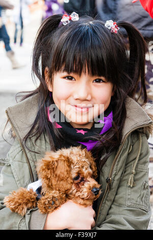 Japanisches Kind, Mädchen, 9-10 Jahre alt, in Mantel, lächeln und posieren für Viewer während sie Streicheleinheiten und hält in beiden Armen ihr Haustier Hund. Auge - Kontakt. Stockfoto