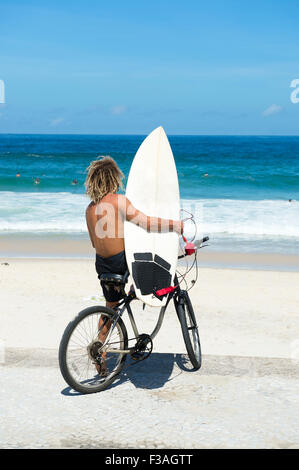 Brasilianische Surfer auf einem Fahrrad sitzen auf Fahrrad mit Blick auf die Wellen Ipanema Strand Rio de Janeiro Brasilien Stockfoto