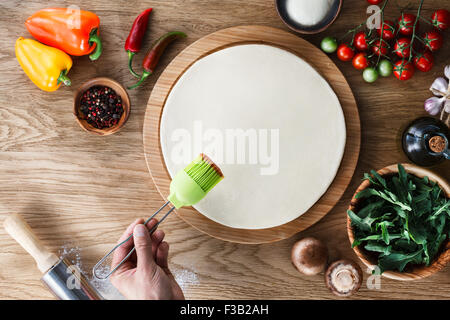 Kochprozess Pizza: Tomatensauce auf einen Pizzateig mit einem Silikon-Gebäck-Pinsel zu verbreiten. Hölzerne Hintergrund, Ansicht von oben. Stockfoto