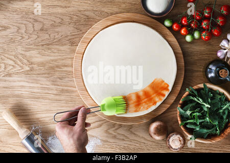 Kochprozess Pizza: Tomatensauce auf einen Pizzateig mit einem Silikon-Gebäck-Pinsel zu verbreiten. Hölzerne Hintergrund, Ansicht von oben. Stockfoto