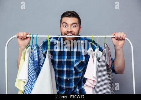 Porträt eines Mannes glücklich Hipster mit Rack von Kleidung auf grauem Hintergrund stehend und Blick in die Kamera Stockfoto