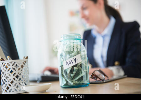 Frau im Büro mit Glas voller Geld auf ihrem Schreibtisch Stockfoto