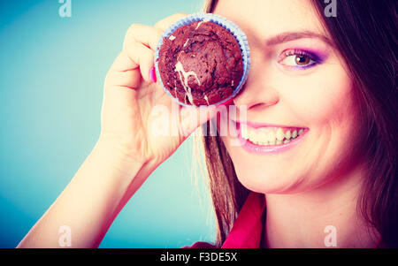 Diät süß Essen und Leute-Konzept. Lustige Frau hält Kuchen in der hand Spaß über ihr Auge mit Cupcake blauen Hintergrund Stockfoto