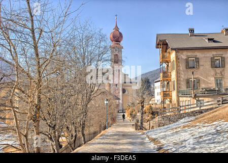 Der Luis Trenker Promenade führt bis St. Ulrich, italienisches Dorf in Dolomiten Alpen: typische Häuser, Gebäude und die roten Glockenturm mit Uhr Pfarrkirche St. Ulrich mit schneebedeckten Bergen und grünen Koniferen im Hintergrund Stockfoto