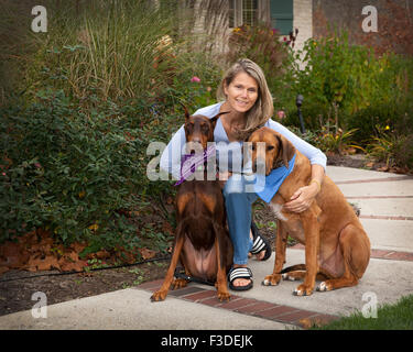 Applying attraktive Frau auf Bürgersteig mit den 2 großen Hund Gefährtinnen. Ein Raum des grünen Laub, Pflanzen zu ihrer linken Seite. Stockfoto