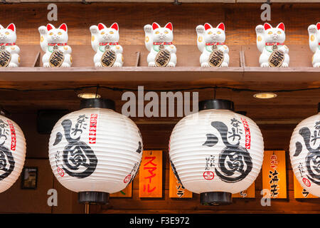 Osaka - Shinsekai, Maneki Neko, Reihe von Beckoning Cat (viel Glück) zahlen im Regal mit chochin, Papier Laternen, unten Restaurant im freien Hängen. Stockfoto