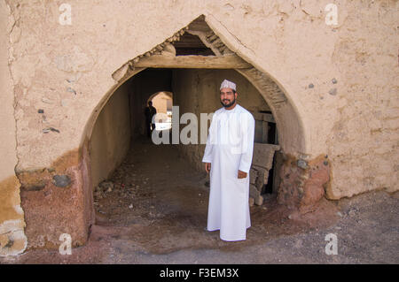 Omanische Mann in traditionellen Dishdasha Robe steht vor einem uralten Adobe Torbogen mit einem anderen Mann im Schatten im Hintergrund Stockfoto