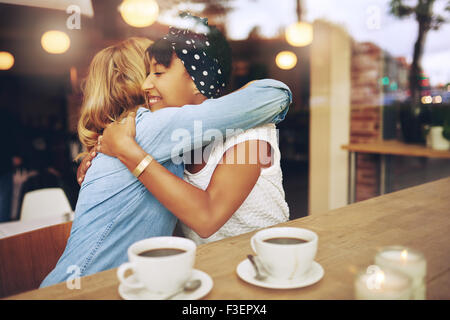 Zwei Multi ethnischen zärtlich Freundinnen umarmen, wie sie sitzen in einem Café bei einer Tasse Kaffee zusammen Stockfoto