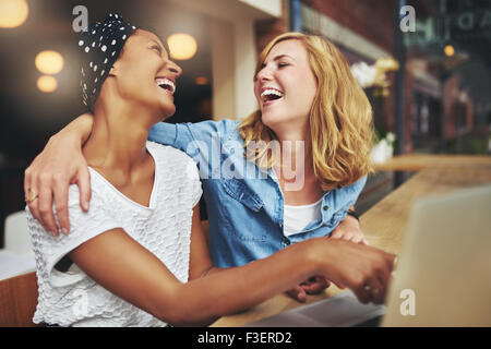 Zwei liebevolle multirassische Freundinnen umarmen und lachen, als sie auf einem Laptop-Computer-Bildschirm zeigen, dass sie teilen Stockfoto