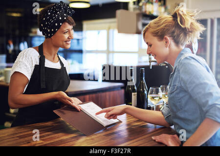 Kundin wählen Sie Wein aus einer Weinkarte präsentiert ihr durch eine charmante junge afrikanische amerikanische Barkeeper in einer Bar co Stockfoto