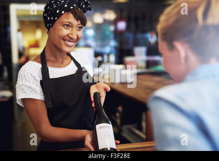 Lächelnd attraktive junge afrikanische amerikanische Kellnerin oder Barkeeper zeigt eine Flasche Rotwein zu einem Kunden in einer Bar vor übe Stockfoto