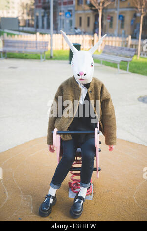 junge schöne asiatische Hipster Frau sitzt auf einem Spielzeug ein Spielgelände mit Kaninchen-Maske - Karneval, Halloween, verrückten Konzept Stockfoto