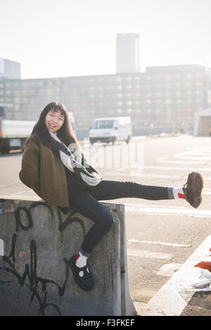 junge schöne asiatische Hipster lange braune glatte Haare Frau sitzt auf einer kleinen Mauer, Spaß haben, suchen Sie in der Kamera lachen, während im Sonnenuntergang bei Gegenlicht - Sorglosigkeit, Jugendkonzept Stockfoto