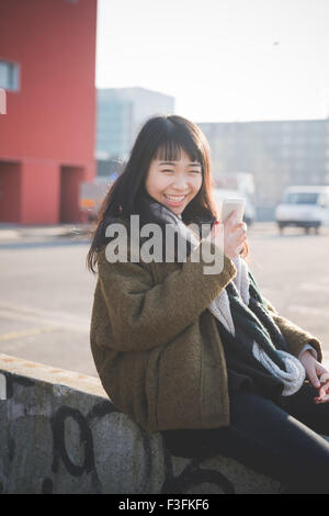 junge schöne asiatische Hipster lange braune glatte Haare Frau sitzt auf einer Mauer mit einem Smartphone suchen in der Kamera, lachen, während des Sonnenuntergangs bei Gegenlicht - Technologie, soziale Netzwerk-Konzept Stockfoto