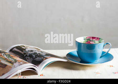 Ein Magazin über eine florale Tisch mit einem blauen Tasse Tee auf einer Untertasse vor einer weißen Wand Stockfoto