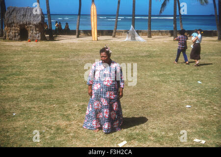 Touristen besuchen eine Luau auf Hawaii in den 1950er Jahren mit Unterhaltung von Hawaiianer. Frau in traditioneller Kleidung mit Surfbrett aufgestützt neben Palmen Stockfoto