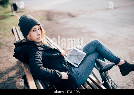 junge schöne Hipster sportliche blonde Frau sitzen auf einer Bank in einem Park mit Fahrrad und Tablet, auf der Suche in der Kamera gelehnt ihre Beine auf dem Rad - Musik, sportliche Konzept Stockfoto