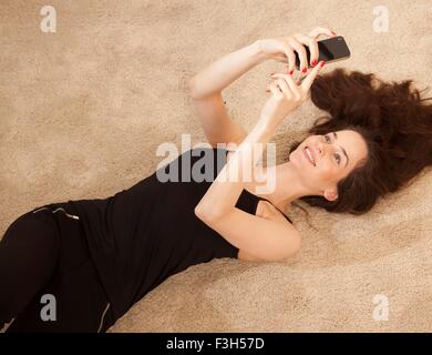 Erhöhte Ansicht der jungen Frau liegt auf Teppich mit Smartphone lächelnd Stockfoto