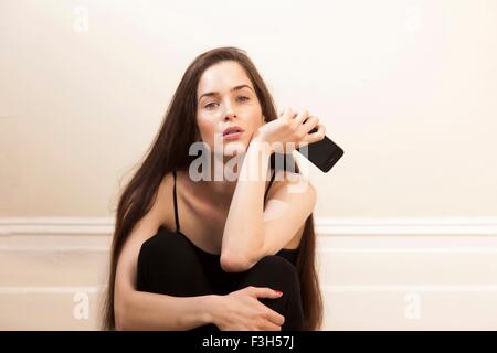 Junge Frau mit Smartphone sitzen umarmt Knien Blick in die Kamera Stockfoto