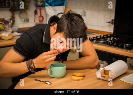 Attraktive junge Mann Essen Frühstück, mit Ellenbogen stützte sich auf Küchentisch und halten Tasse Kaffee zum Frühstück Stockfoto