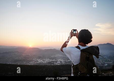 Rückansicht des jungen Mannes Fotografieren von Landschaft und Sonnenuntergang auf Smartphone, Javea, Spanien Stockfoto