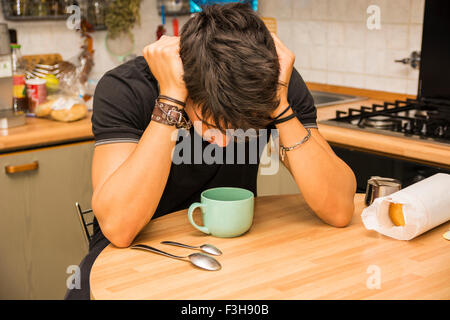 Müde oder krank attraktiver junger Mann mit geschlossenen Augen und Hand in den Haaren, mit Ellenbogen stützte sich auf Küchentisch und halten Tasse Cof Stockfoto