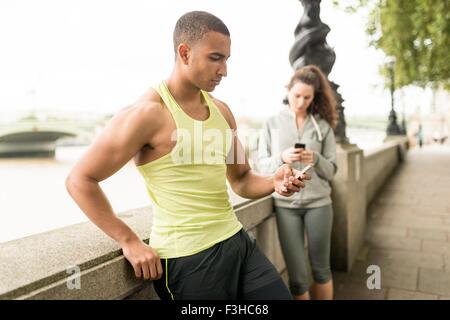 Männliche und weibliche Läufer SMS auf Smartphone im riverside Stockfoto