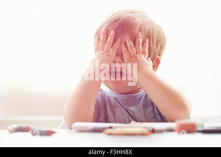 Junge sitzt am Tisch ruht auf Ellbogen, Mund öffnen, für Augen mit Händen Stockfoto